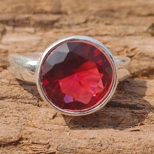 Può includere: Un anello d'argento con una grande gemma rotonda, sfaccettata, di colore rosso intenso. L'anello è posto su una superficie di legno marrone strutturata, con la gemma che cattura la luce. La fascia dell'anello è semplice e liscia.