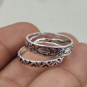 May include: Two silver rings with detailed patterns. The top ring has a floral design, while the bottom ring features a repeating heart pattern. The rings are displayed on a hand, highlighting their intricate craftsmanship. The rings are likely to be worn as jewellery.