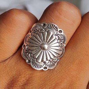 Op de afbeelding: Een zilverkleurige ring met een gedetailleerd bloemmotief. De ring heeft een centraal zonnestraalpatroon omgeven door sierlijke details en kleine cirkelvormige accenten. De ring wordt aan een vinger gedragen en toont zijn ingewikkelde vakmanschap.
