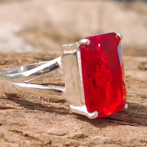 May include: A silver ring with a large, rectangular, bright red gemstone. The ring is set on a wooden surface, with a blurred blue object in the background. The gemstone is the focal point, showcasing its vibrant color and cut.