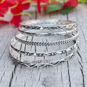 May include: A stack of silver bangles with intricate floral and leaf patterns. The bangles are displayed on a weathered wooden surface, with red flowers in the background. The bangles have various designs, adding a touch of elegance.