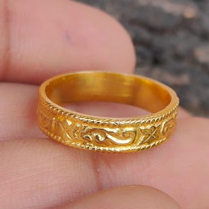 Peut inclure: Une bague dorée ornée d'un motif complexe de volutes et de motifs floraux. La bague est tenue dans une main, avec un arrière-plan flou et texturé. La bague semble être en métal.