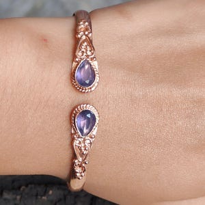 May include: Rose gold cuff bracelet with two teardrop-shaped, purple gemstones. The open design bracelet has ornate detailing around the gemstones. The bracelet is worn on a wrist.