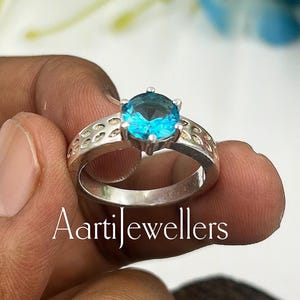 May include: A silver ring with a round, bright blue gemstone. The band has a decorative pattern. The text "Aartijewellers" is visible on the ring.