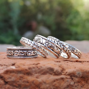 Puede incluir: Cuatro anillos de plata con intrincados diseños florales y trenzados, expuestos sobre una superficie texturizada de color marrón rojizo. Los anillos varían en anchura y detalle, mostrando una gama de patrones en relieve. El fondo está borroso, con toques de follaje verde.