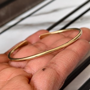 May include: A gold-colored, open cuff bracelet. The bracelet is a simple, minimalist design with a smooth, polished finish. The bracelet is held in a hand, with a marble and black background.