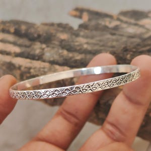 Könnte beinhalten: Ein silbernes Armreif-Armband mit einem komplizierten, sich wiederholenden Muster. Das runde Armband scheint aus Metall zu sein. Das detaillierte Design bedeckt einen Teil der Oberfläche des Armbands und verleiht dem Schmuck optisches Interesse.