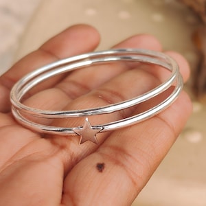 Peut inclure: Un bracelet jonc double en argent doté d'une breloque en forme d'étoile à cinq branches fixée sur l'anneau inférieur. Le bracelet présente une finition métallique polie et réfléchissante ainsi qu'un profil lisse et arrondi.