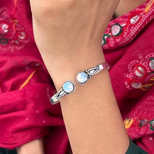 Könnte beinhalten: Ein silberfarbenes Armreif-Armband mit zwei runden, irisierenden Mondstein-Cabochons. Das offene Armband hat dekorative Details in der Nähe der Steine. Der Hintergrund zeigt ein rotes Kleidungsstück mit Stickereien.