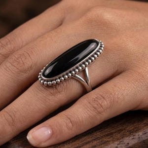 May include: A silver ring with a large, oval black stone. The stone is framed by a beaded edge. The ring's band has a split design. The ring is displayed on a hand.