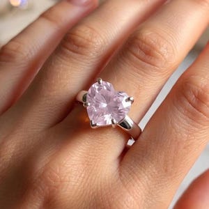 May include: A silver ring featuring a large, heart-shaped, light pink gemstone. The ring is set with four prongs and is worn on a finger. The background is blurred, with hints of other jewelry and soft lighting.