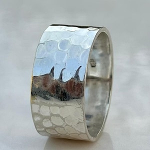 Puede incluir: Un anillo ancho de plata con textura martillada. El anillo tiene un interior liso y pulido y un exterior texturizado. El diseño martillado crea un patrón de pequeñas hendiduras irregulares en la superficie. El anillo está sobre una superficie blanca.