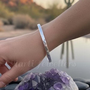 Op de afbeelding: Een zilverkleurige metalen armband met een gehamerde textuur. De armband heeft een glad, reflecterend oppervlak en een eenvoudig, open ontwerp. Het wordt om de pols gedragen en toont de metalen afwerking en subtiele, onregelmatige inkepingen.