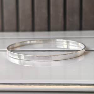 May include: A silver bangle bracelet with a flat, polished surface. The bracelet is circular and reflects light, showcasing a minimalist design. The jewelry is displayed on a white surface.