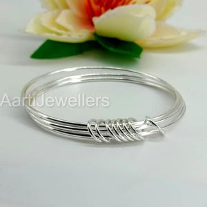 May include: A stack of silver bangles with a coiled design at the center. The bangles are thin and round, with a bright, reflective surface. The text "AartiJewellers" is visible on the left side of the image. A flower is in the background.