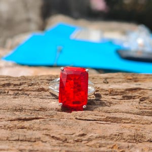 May include: A silver ring featuring a large, rectangular, vibrant red gemstone. The ring is set on a textured, brown wooden surface, with a blurred blue background.