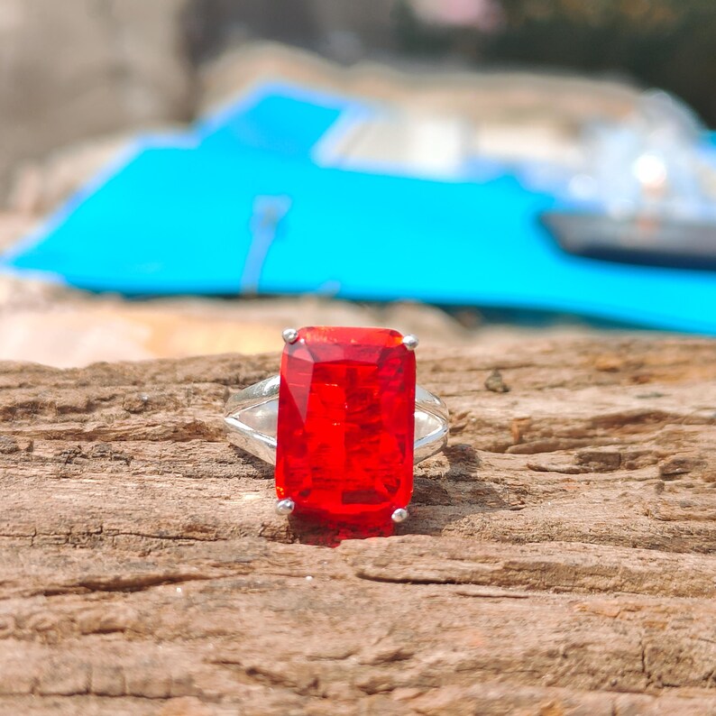 May include: A silver ring featuring a large, rectangular, faceted, bright red gemstone. The ring is set on a wooden surface, with the gemstone prominently displayed. The ring's band is a simple, elegant design.