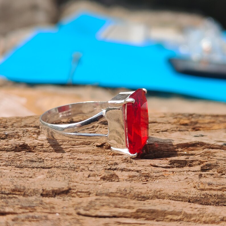 May include: A silver ring featuring a large, rectangular, faceted red gemstone. The ring is set on a wooden surface with a blurred blue background. The gemstone is the focal point, catching the light and showcasing its vibrant color.
