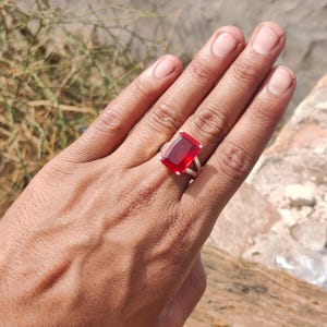 May include: A silver ring with a large, rectangular, deep red gemstone. The ring is set on a hand, showcasing the vibrant color and design. The gemstone is the focal point, catching the light and drawing attention.