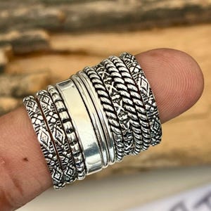 May include: A collection of silver rings with various designs, including braided, patterned, and smooth bands. The rings are stacked together, showcasing their intricate details and textures. The rings are displayed on a finger.