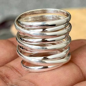 May include: A multi-band ring crafted from polished silver, featuring several interconnected, smooth, rounded wire loops that wrap around to create a wide, stacked appearance with a reflective, metallic finish throughout the entire piece.
