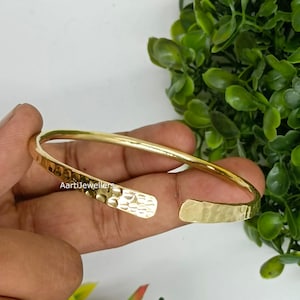 May include: A gold-toned open cuff bracelet featuring a hammered, textured surface throughout the band. The metal has a reflective finish, and the text 'AartiJewellers' is clearly visible on the inner side of the bracelet's curved metal surface.
