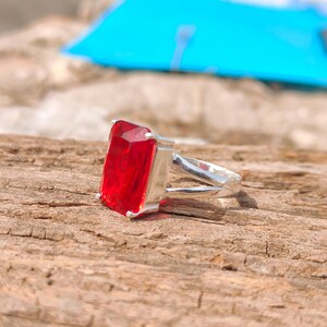 May include: A silver ring featuring a large, rectangular, faceted red gemstone. The ring's band splits into two parallel strands as it approaches the gemstone, creating a unique design. The ring is set against a textured wooden surface.