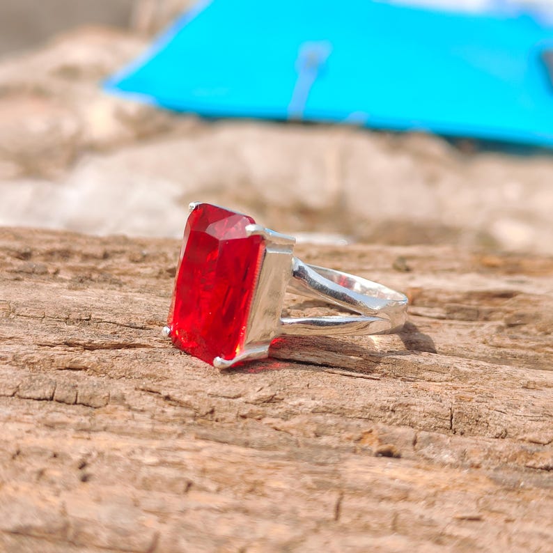 May include: A silver ring featuring a large, rectangular, faceted red gemstone. The ring's band splits into two parallel strands as it approaches the gemstone, creating a unique design. The ring is set against a textured wooden surface.