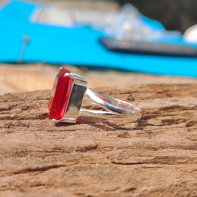 May include: A silver ring with a large, rectangular, red gemstone. The gemstone is the focal point, set in a silver band. The ring is displayed on a wooden surface, with a blurred blue background.