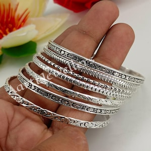 Op de afbeelding: Een verzameling zilveren armbanden met ingewikkelde bloemen- en geometrische patronen. De armbanden zijn gestapeld en tonen verschillende ontwerpen, waaronder bladeren, kralen en gedetailleerde randen. De sieraden worden op een hand getoond, wat de vakmanschap en de reflecterende kwaliteit van het metaal benadrukt.