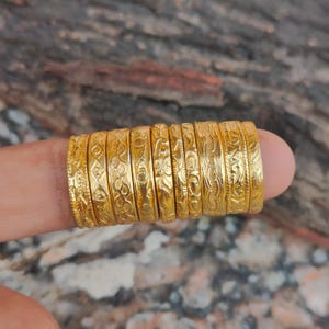 Pode incluir: Uma coleção de anéis dourados, cada um com um padrão gravado único. Os anéis estão empilhados em um dedo, mostrando a variedade de designs. O fundo é uma superfície texturizada desfocada.