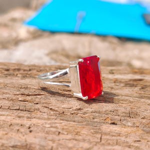 May include: A silver ring featuring a large, rectangular, vibrant red gemstone. The ring's band has a split design, and the gemstone is set in a square silver setting. The ring is displayed on a textured, brown wooden surface.