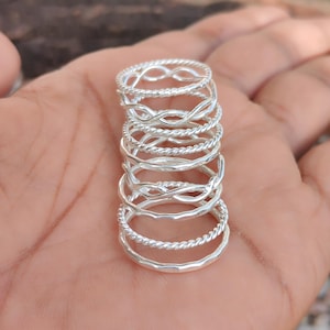 May include: A stack of seven silver-toned rings featuring various textures and designs, including twisted rope patterns, smooth bands, and infinity-style links, all arranged vertically in a neat, uniform column of metallic jewelry.