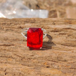 May include: A silver ring featuring a large, rectangular, faceted red gemstone. The ring's band is a simple, polished silver, and the gemstone is the focal point, catching the light and displaying a vibrant red color. The ring is set against a natural wood background.