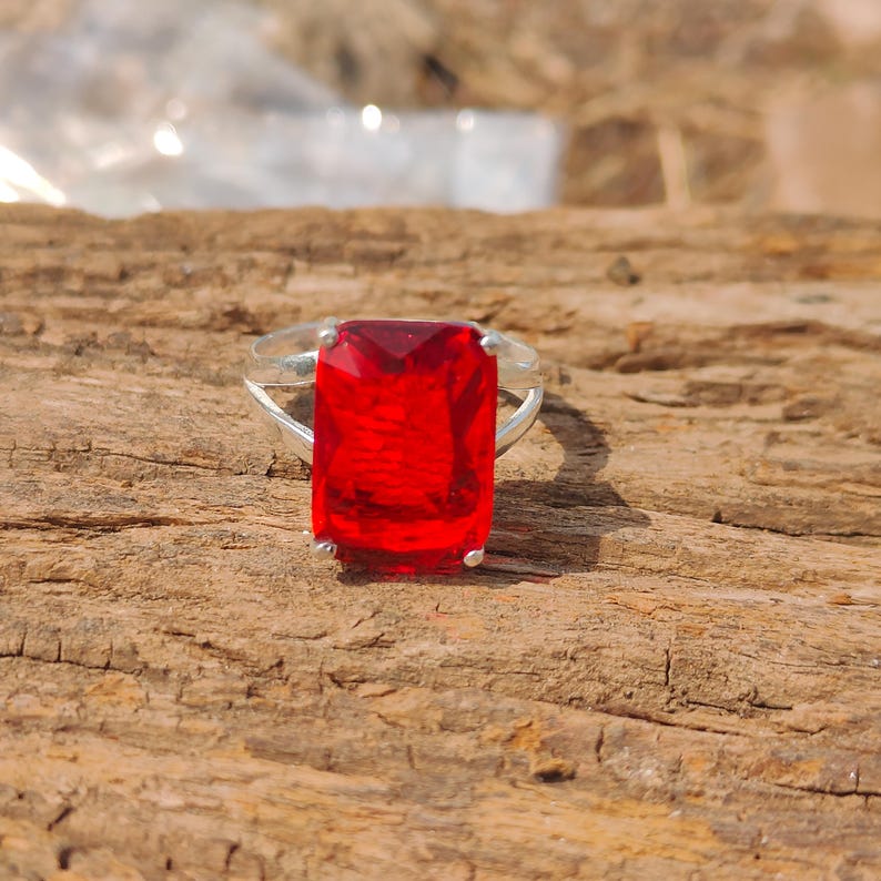 May include: A silver ring featuring a large, rectangular, faceted red gemstone. The ring's band is a simple, polished silver, and the gemstone is the focal point, catching the light and displaying a vibrant red color. The ring is set against a natural wood background.