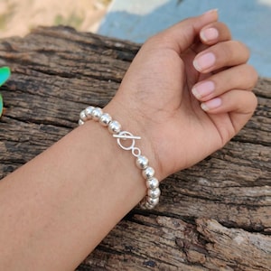 Könnte beinhalten: Ein silberfarbenes Perlenarmband bestehend aus einer Reihe gleichmäßiger, polierter, kugelförmiger Perlen. Das Armband wird mit einem Knebelverschluss geschlossen, der aus einem kreisförmigen Ring und einem Querstab in passendem Silberton besteht.