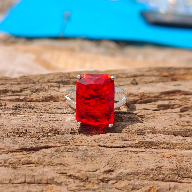 May include: A silver ring featuring a large, rectangular, faceted red gemstone. The gemstone is the focal point, set in a simple, elegant silver band. The ring is displayed on a textured, wooden surface.