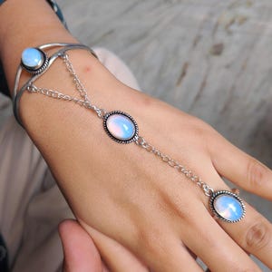 Puede incluir: Una pulsera de cadena de mano plateada con tres piedras ovaladas opalescentes. Una piedra está engastada en la pulsera, otra en el anillo y otra en el centro, conectadas por una delicada cadena. Las piedras tienen una calidad nacarada e iridiscente.