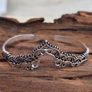 Puede incluir: Un brazalete de metal en tono plateado con un intrincado diseño de filigrana calada y un motivo central de arco apuntado. La superficie del metal tiene un acabado oscuro y oxidado en las áreas hundidas. El texto AartiJewellers es visible.