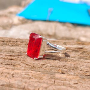 May include: A silver ring featuring a large, rectangular, faceted red gemstone. The ring's band splits into two parallel strands on either side of the gemstone, creating a unique design. The ring is set against a natural wood background.