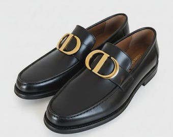 May include: A pair of black leather loafers with a gold-colored metal accent in the shape of a "D" on the vamp. The shoes have a classic design with a rounded toe and a low heel, suitable for various occasions.