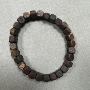 May include: A dark brown wooden bead bracelet. The bracelet is made of square-shaped beads strung together. The beads have a natural wood grain pattern and vary in shades of brown. The bracelet is displayed on a light gray surface.