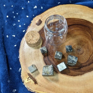 May include: A clear glass jar with a cork lid sits on a wooden slice, surrounded by several small, metallic pyrite cubes. The background is a dark blue fabric with silver flecks.