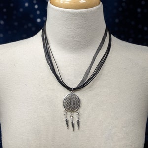 May include: A silver necklace with a circular pendant featuring a geometric design and four dangling feathers. The necklace is on a black ribbon and is displayed on a white mannequin.