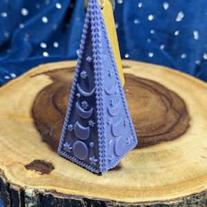 May include: A lavender-colored, pyramid-shaped candle with crescent moon and star designs. The candle has a decorative border and is attached to a wooden tag. The candle is displayed on a wooden slice against a blue background.