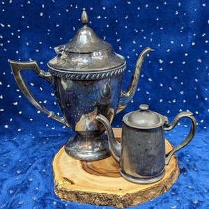 May include: Two silver teapots on a wooden base. The larger teapot has a tall, ornate spout and a round body. The smaller teapot has a shorter spout and a more rounded body. Both teapots have a shiny, polished finish.