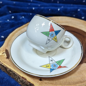 May include: A white teacup and saucer with a gold trim. The cup and saucer feature a colorful five-pointed star with Masonic symbols.