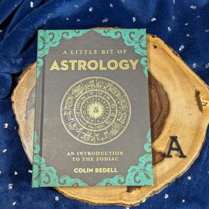 May include: A book titled "A Little Bit of Astrology" with a gold zodiac wheel design on the cover. The book is on a wooden slab, set against a blue background. The book's cover has teal accents.