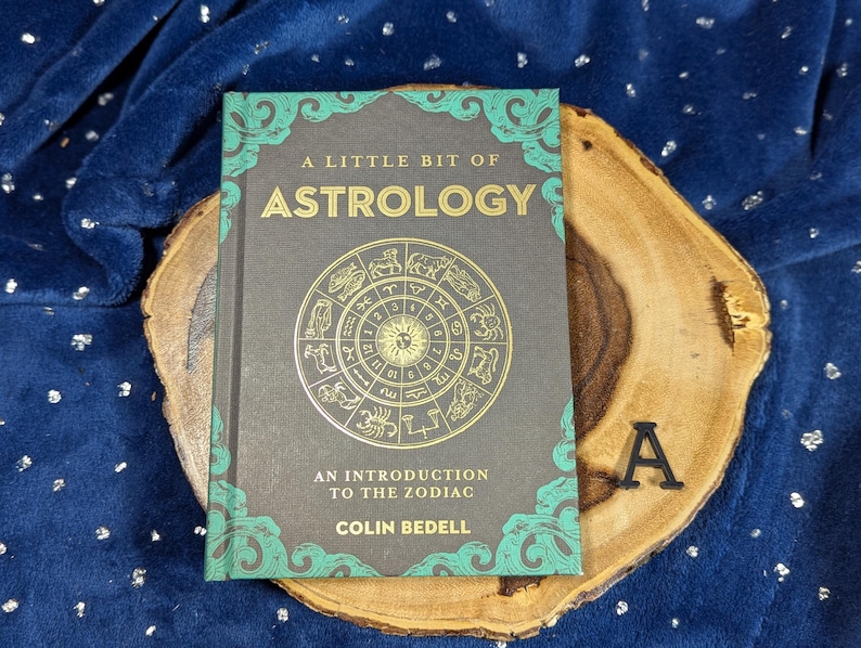 May include: A book titled "A Little Bit of Astrology" with a gold zodiac wheel design on the cover. The book is on a wooden slab, set against a blue background. The book's cover has teal accents.