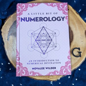 May include: A book titled "A Little Bit of Numerology" with a geometric design on the cover. The book is on a wooden slice with a dark blue background. The author's name is Novalee Wilder.